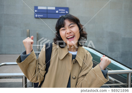 Enthusiastic asian girl stands on street, makes hooray celebration gesture, says yes, triumphing, winning and enjoying this feeling, standing alone outdoors Enthusiastic asian girl stands on street, makes hooray celebration gesture, says yes, triumphing, winning and enjoying this feeling, standing alone outdoors 98921896