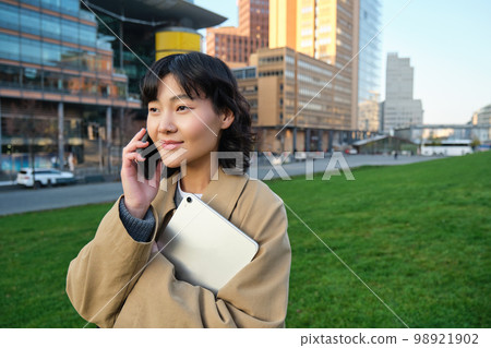 Stylish smiling girl, university student in trench coat, holds tablet, talks on mobile phone, has conversation over telephone and looks relaxed, stands on street Stylish smiling girl, university student in trench coat, holds tablet, talks on mobile phone, has conversation over telephone and looks relaxed, stands on street 98921902