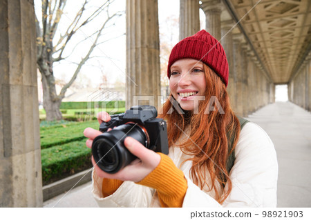 Young redhead female photographer, makes lifestyle shooting in city centre, takes photos and smiles, looks for perfect shot, makes picture Young redhead female photographer, makes lifestyle shooting in city centre, takes photos and smiles, looks for perfect shot, makes picture 98921903