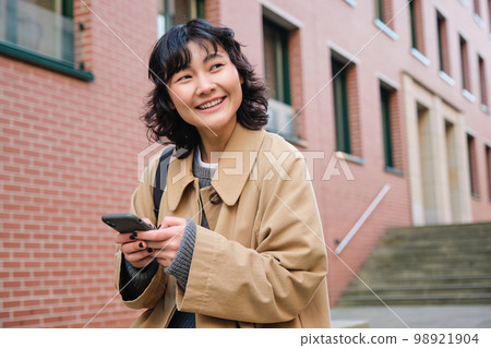 Happy korean girl walks on street, listens music in wireless earphones and holds smartphone, picks song in playlist while standing outdoor near building, reading message Happy korean girl walks on street, listens music in wireless earphones and holds smartphone, picks song in playlist while standing outdoor near building, reading message 98921904