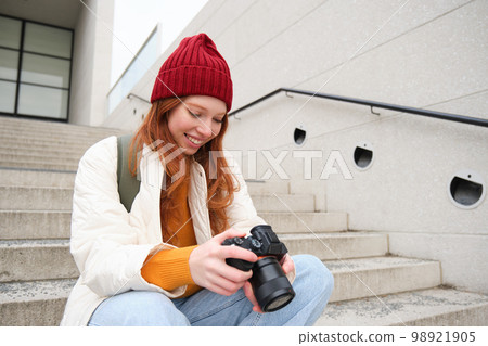 Smiling redhead girl photographer, checks her shots, holds camera and looks at screen, takes photos outdoors, walks around street and does streetstyle shooting Smiling redhead girl photographer, checks her shots, holds camera and looks at screen, takes photos outdoors, walks around street and does streetstyle shooting 98921905