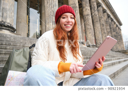 Outdoor shot of young stylish redhead girl sits on staircase and connects to public wifi, uses digital tablet, reads news on gadget 98921934