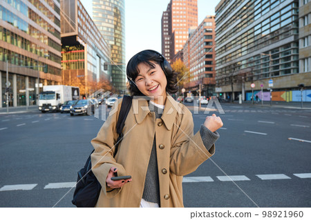 Portrait of young asian girl, student walks in city, listens music in headphones and uses mobile phone on streets, celebrates, triumphs and makes joyful fist pump Portrait of young asian girl, student walks in city, listens music in headphones and uses mobile phone on streets, celebrates, triumphs and makes joyful fist pump 98921960
