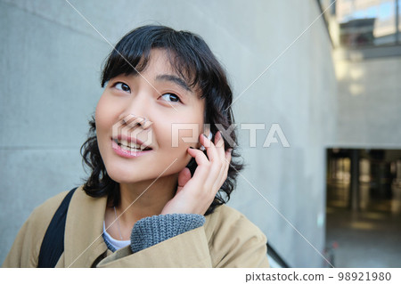 Close up portrait of smiling korean girl in headphones, listens music in wireless earphones, enjoys travelling around town with her favorite songs playlist Close up portrait of smiling korean girl in headphones, listens music in wireless earphones, enjoys travelling around town with her favorite songs playlist 98921980