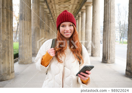 Happy young female model stands on street and holds mobile phone, uses smartphone app, follows the route of online map 98922024