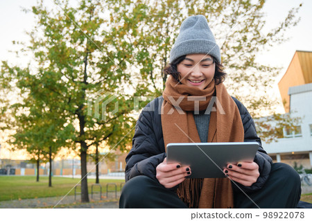 Portrait of asian girl in warm clothes, sits on bench with digital tablet and graphic pen, smiling happily, draws outdoors in chilly weather Portrait of asian girl in warm clothes, sits on bench with digital tablet and graphic pen, smiling happily, draws outdoors in chilly weather 98922078