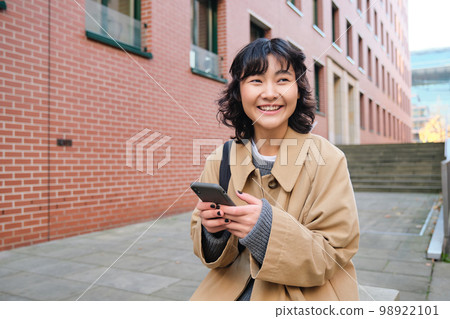Stylish korean hipster girl, walks in city centre, sends text message, uses location map application on smartphone while walking down the street, wears trench coat Stylish korean hipster girl, walks in city centre, sends text message, uses location map application on smartphone while walking down the street, wears trench coat 98922101