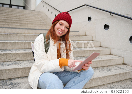 Happy stylish redhead girl, student in red hat, holds digital tablet, uses social media app, searches something online, connects to wifi Happy stylish redhead girl, student in red hat, holds digital tablet, uses social media app, searches something online, connects to wifi 98922103