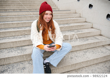 Hipster ginger girl, redhead woman sits on stairs with smartphone, waits for someone and messages on social media on mobile phone app Hipster ginger girl, redhead woman sits on stairs with smartphone, waits for someone and messages on social media on mobile phone app 98922124
