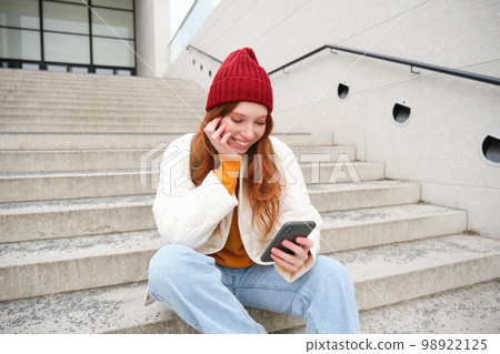Beautiful redhead teen girl, sits on stairs and smiles, holds mobile phone, uses smartphone application, chats on telephone app Beautiful redhead teen girl, sits on stairs and smiles, holds mobile phone, uses smartphone application, chats on telephone app 98922125