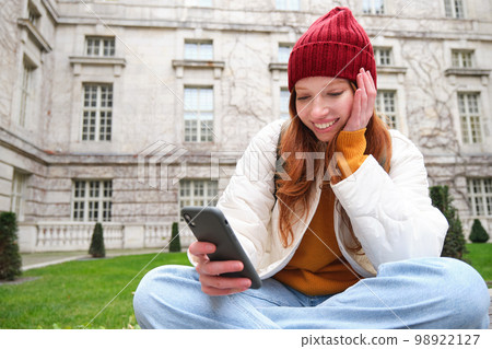 Portrait of stylish young woman, 25 years, sits on bench in park and uses mobile phone, reads online news, messages or watches video on smartphone app, connects to public wifi Portrait of stylish young woman, 25 years, sits on bench in park and uses mobile phone, reads online news, messages or watches video on smartphone app, connects to public wifi 98922127