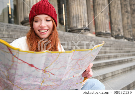 Smiling girl traveler, tourist sits on stairs near museum and holds paper map, explores city during sightseeing adventure trip Smiling girl traveler, tourist sits on stairs near museum and holds paper map, explores city during sightseeing adventure trip 98922129