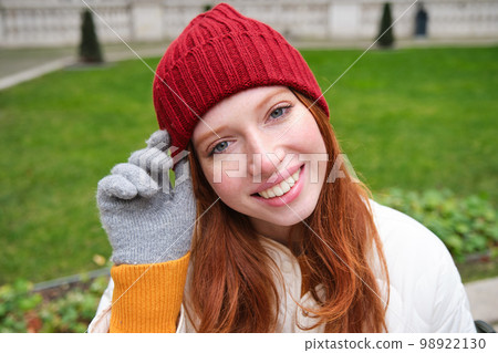 Close up portrait of beautiful redhead woman in red knitted hat, warm gloves, smiling and looking happy at camera, sitting in park Close up portrait of beautiful redhead woman in red knitted hat, warm gloves, smiling and looking happy at camera, sitting in park 98922130