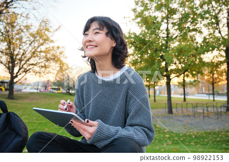 Young korean woman sits in park, draws on digital tablet, writes in gadget with pen tool, looks around and makes notes, creates artwork or scatches 98922153