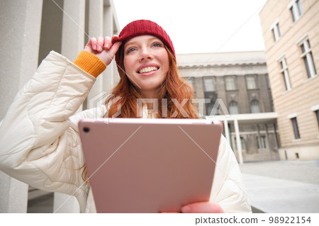 Happy redhead girl in red hat, walks around city with digital tablet, connects to public internet wifi and looks for route, looks at map on her gadget Happy redhead girl in red hat, walks around city with digital tablet, connects to public internet wifi and looks for route, looks at map on her gadget 98922154