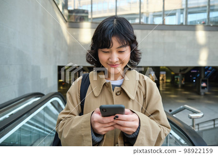People in city. Young korean woman travels around city, goes up the escalator, uses her mobile phone and smiles People in city. Young korean woman travels around city, goes up the escalator, uses her mobile phone and smiles 98922159