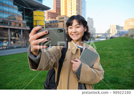 Happy university student, korean girl takes selfie with her papers and digital tablet, holds smartphone and poses near university campus Happy university student, korean girl takes selfie with her papers and digital tablet, holds smartphone and poses near university campus 98922180