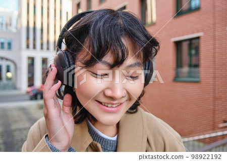 Close up portrait of stylish brunette asian girl, listens music in headphones, touches earphones and smiles, enjoys favorite song, stands on street Close up portrait of stylish brunette asian girl, listens music in headphones, touches earphones and smiles, enjoys favorite song, stands on street 98922192