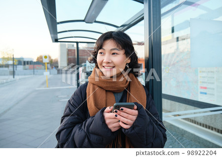 Portrait of Korean woman in winter jacket, standing with smartphone, waiting for bus on stop, looking at mobile phone app checking public transport application Portrait of Korean woman in winter jacket, standing with smartphone, waiting for bus on stop, looking at mobile phone app checking public transport application 98922204