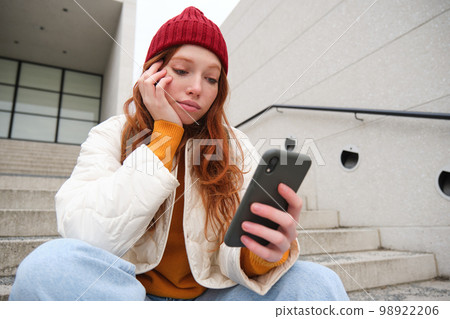 Mobile phones and people. Young stylish redhead girl sits on stairs with telephone, uses smartphone app, reads smth online 98922206