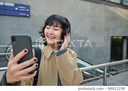 Beautiful korean girl in headphones, takes selfie on her smartphone, stands on street with mobile phone, records video, listens music and travels around city Beautiful korean girl in headphones, takes selfie on her smartphone, stands on street with mobile phone, records video, listens music and travels around city 98922298