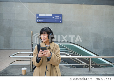 Portrait of stylish asian hipster girl, listens music in headphones, looks at her phone, tourist looking at map for a way to sightseeing, drinking coffee to go 98922333
