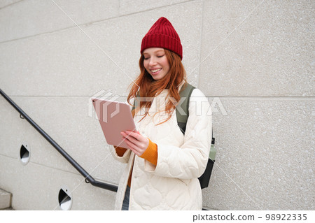 Young redhead woman with red hat, uses her digital tablet outdoors, stands on street with gadget, connects to wifi internet and searches for a location in internet 98922335