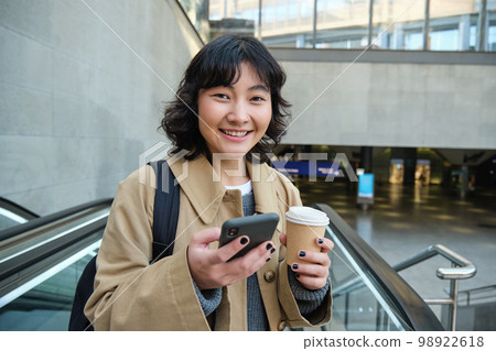 Portrait of beautiful brunette girl on escalator, drinks coffee to go and uses smartphone, smiles at camera, goes to work or univeristy, commutes 98922618