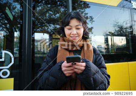 Beautiful korean girl, student on bus stop, looking at her smartphone, checking timetable, reading text message, wearing winter clothes 98922695