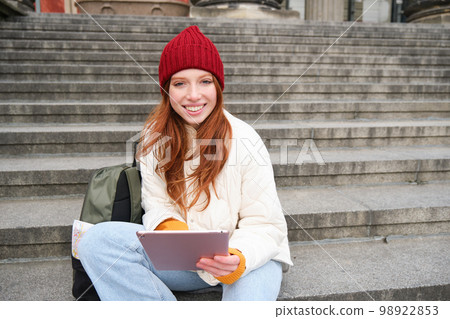 Beautiful young modern girl with red hair, holds digital tablet, sits on stairs near museum and connects public internet, sends message on gadget app 98922853