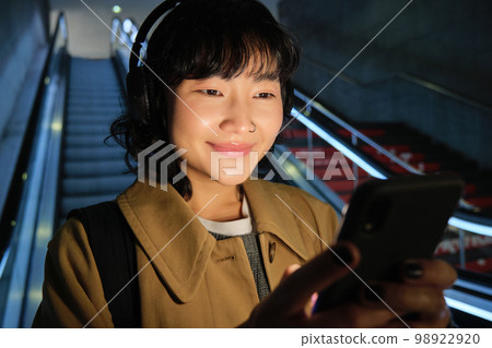 Close up of cute asian girl in headphones, picks song to listen while commuting, going down escalator to metro, smiling as reading message on mobile phone 98922920