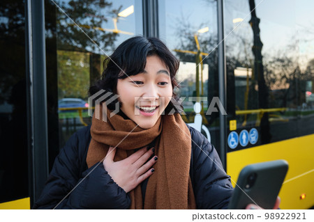Portrait of cheerful asian girl talks on mobile phone, video chats, looks amazed at smartphone camera, stands on bus stop 98922921