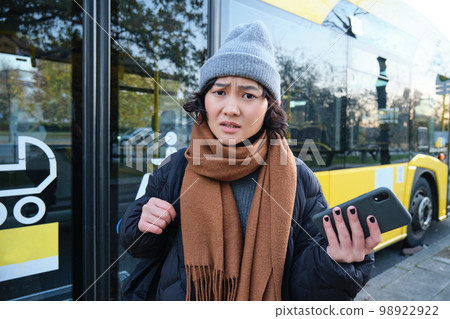 Portrait of confused asian girl, standing on bus stop, holding mobile phone, looking shocked and upset, disappointed by public transport delay 98922922