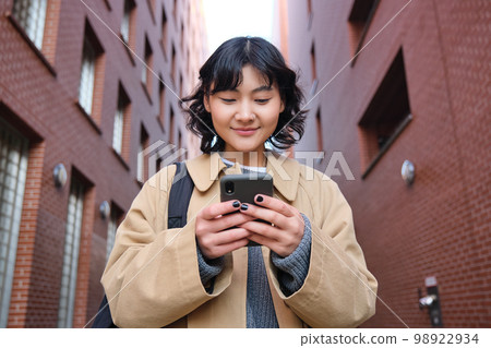 Lower angle view of brunette korean girl, listening music in headphones, walking along street and looking at smartphone, reading message on mobile phone Lower angle view of brunette korean girl, listening music in headphones, walking along street and looking at smartphone, reading message on mobile phone 98922934