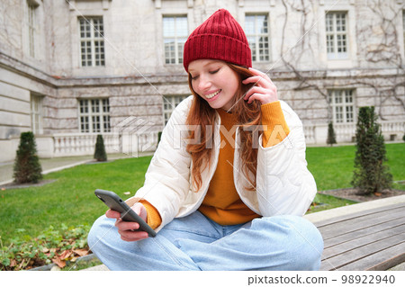 Young smiling redhead girl sits on bench and uses smartphone app, reads news online, watches video on mobile phone while relaxed in park 98922940