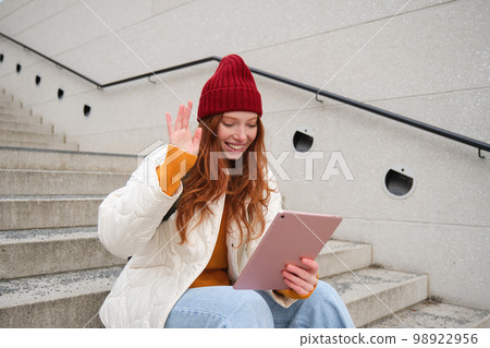 Joyful redhead teen girl, student with digital tablet, says hello, waves hand at gadget camera, connects to video chat, talks to friend in application 98922956