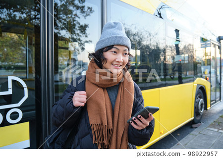 Portrait of girl standing near bus on a stop, waiting for her public transport, schecks schedule on smartphone application, holds mobile phone, wears warm clothes 98922957