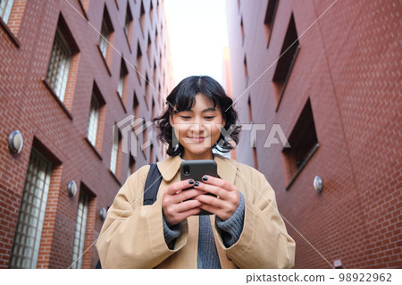 Lower angle view of brunette korean girl, listening music in headphones, walking along street and looking at smartphone, reading message on mobile phone Lower angle view of brunette korean girl, listening music in headphones, walking along street and looking at smartphone, reading message on mobile phone 98922962
