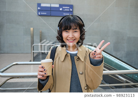 Positive korean girl in headphones, drinks coffee to go, shows peace, v-sign, stands on street and smiles happily 98923113