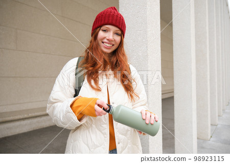 Beautiful redhead woman drinking hot tea or coffee from thermos, female tourist enjoys warm drink, rests during her journey around town 98923115