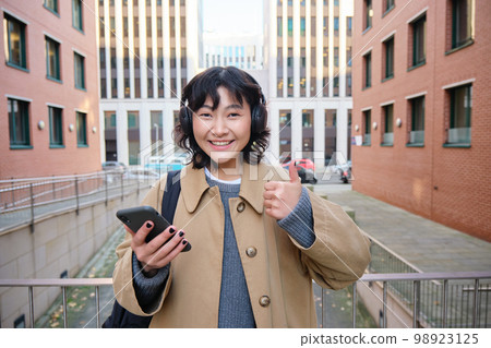 Portrait of cheerful asian girl in headphones, stands on street and shows thumbs up, found something online on her smartphone, using mobile phone 98923125