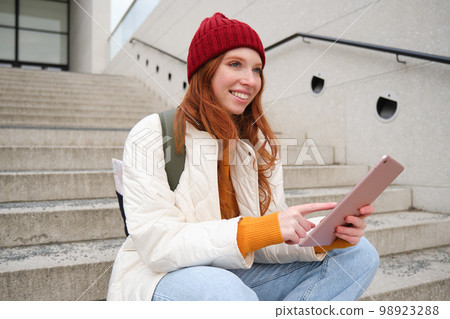 Happy stylish redhead girl, student in red hat, holds digital tablet, uses social media app, searches something online, connects to wifi Happy stylish redhead girl, student in red hat, holds digital tablet, uses social media app, searches something online, connects to wifi 98923288