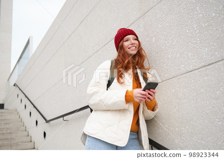 Happy girl student in red hat, holds smartphone, tourist looks at map app on her phone, explores sightseeing, texts message, looks for couchsurfing, rents place to stay online 98923344