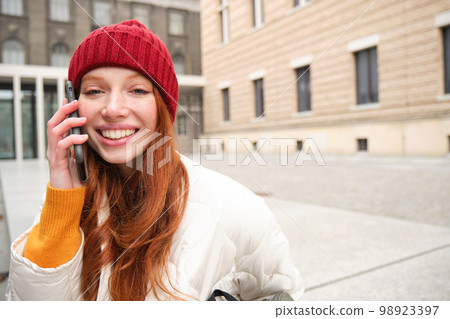 Stylish modern redhead girl talks on mobile phone, makes a telephone call, calling someone on smartphone app from outside, stands on street and smiles 98923397