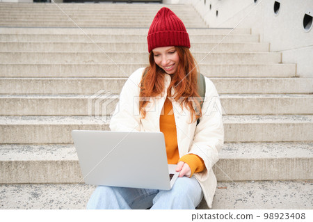 Smiling redhead girl, young woman typing on laptop keyboard, sitting outdoors on stairs with computer, working remote, doing her homework on fresh air Smiling redhead girl, young woman typing on laptop keyboard, sitting outdoors on stairs with computer, working remote, doing her homework on fresh air 98923408