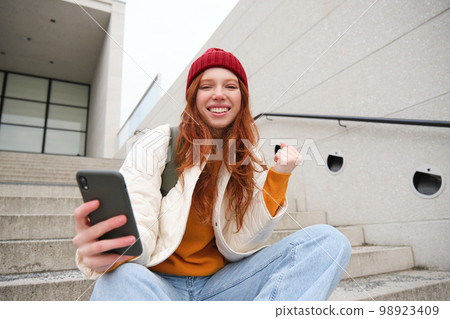 Joyful young redhead girl sits on street stairs, say yes, wins and celebrates with phone in hand, receive positive news on her mobile application and rejoice 98923409