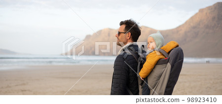 Father rising hands to the sky while enjoying pure nature carrying his infant baby boy son in backpack on windy sandy beach of Famara, Lanzarote island, Spain at sunset. Family travel concept Father rising hands to the sky while enjoying pure nature carrying his infant baby boy son in backpack on windy sandy beach of Famara, Lanzarote island, Spain at sunset. Family travel concept 98923410