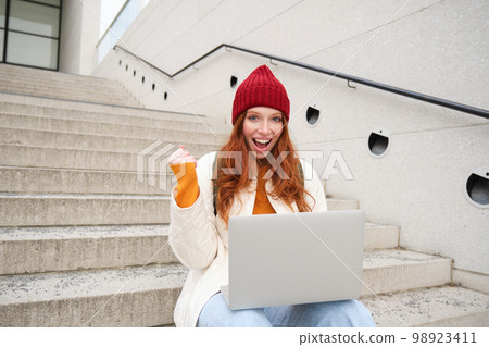 Smiling redhead girl, young woman typing on laptop keyboard, sitting outdoors on stairs with computer, working remote, doing her homework on fresh air Smiling redhead girl, young woman typing on laptop keyboard, sitting outdoors on stairs with computer, working remote, doing her homework on fresh air 98923411