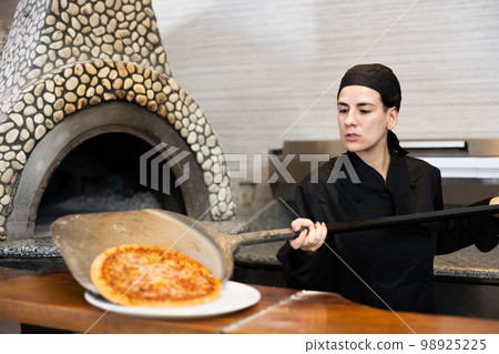Female chef in black uniform prepares pizza and pulls it out of oven in kitchen of pizzeria Female chef in black uniform prepares pizza and pulls it out of oven in kitchen of pizzeria 98925225