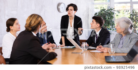 Young Latin female turning on presentation on projector while office coworkers of different ages sitting at table with laptops and notes 98925496